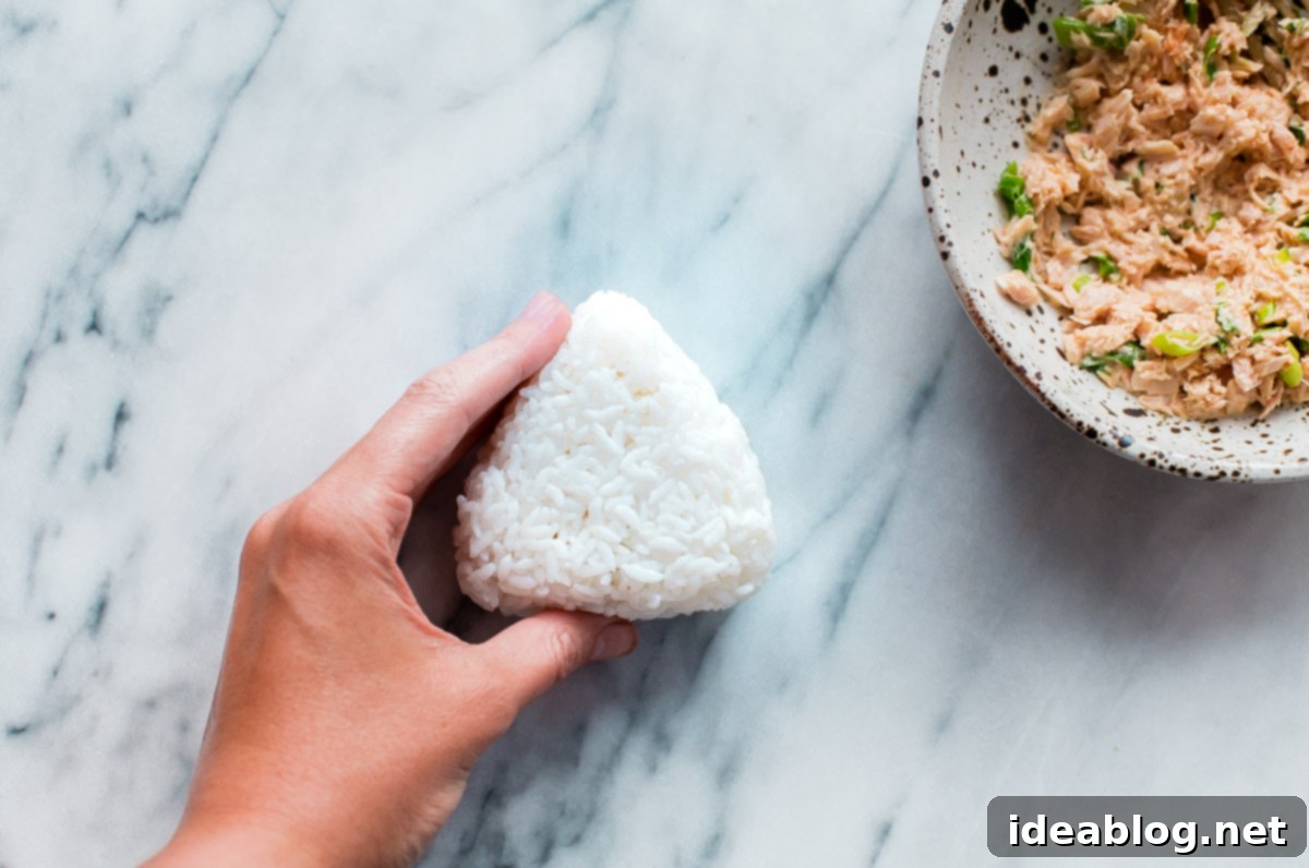 Homemade Spicy Tuna Onigiri - learn how to make homemade nigiri with this simple recipe! This spicy tuna onigiri version is filled with a simple filling of canned olive oil packed tuna, mayonnaise, sriracha, rice vinegar, and scallion. #onigiri #japanese #abeautifulplate #tuna #recipe #snack #glutenfree Step-by-step guide on how to shape Japanese Onigiri by hand, enclosing a filling within perfectly cooked rice.