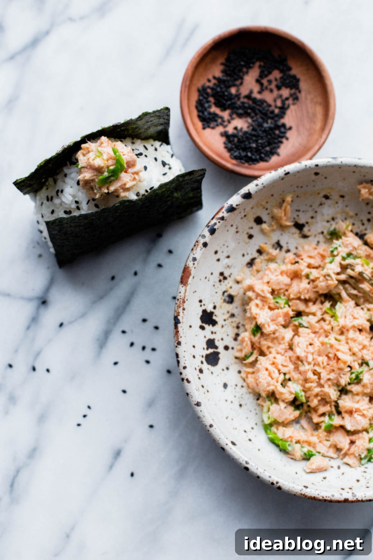 Homemade Spicy Tuna Onigiri - learn how to make homemade nigiri with this simple recipe! This spicy tuna onigiri version is filled with a simple filling of canned olive oil packed tuna, mayonnaise, sriracha, rice vinegar, and scallion. #onigiri #japanese #abeautifulplate #tuna #recipe #snack #glutenfree Beautifully shaped Spicy Tuna Onigiri, garnished with black sesame seeds and wrapped in crisp nori, presented on a wooden board.