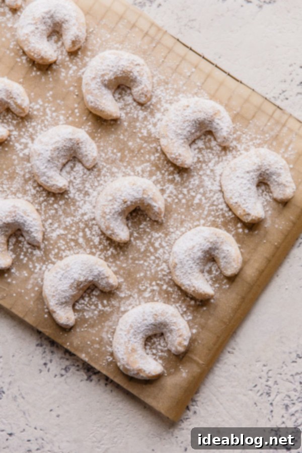 Almond Crescent Cookies - these traditional Almond Crescent Cookies (or Austrian kipferl cookies) are my go-to easy Christmas cookies. Buttery, crisp, and impossible to resist! #abeautifuplate #almondflour #kipferl #almondcrescentcookies #recipe #dessert #Christmas #holiday #baking A hand gently dusting baked almond crescent cookies with powdered sugar.