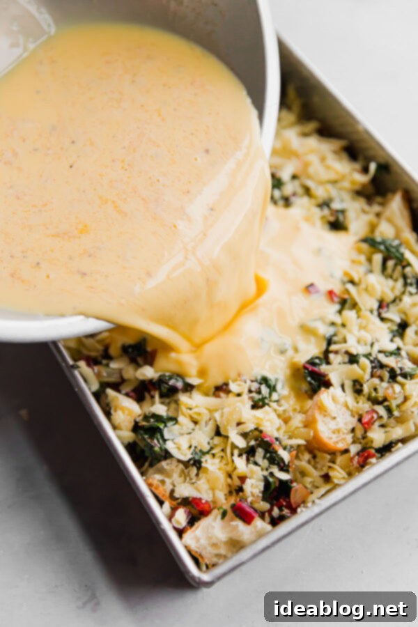 Overnight Vegetarian Breakfast Bake 3 A large bowl of whisked egg and milk mixture being poured evenly over layers of bread, chard, and cheese in a baking dish.